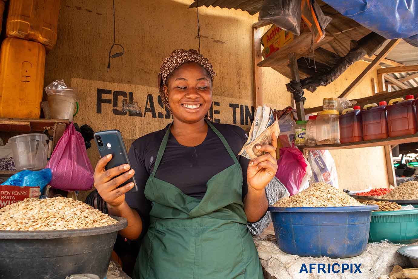 Happy Market Woman Holding Naira Note and POS Machine in Front of Her Shop